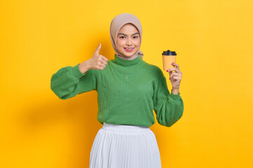 Fototapeta na wymiar Smiling beautiful Asian woman in green sweater holding paper coffee cup, showing thumbs up gesture isolated over yellow background