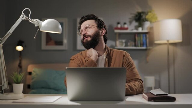 Neck pain, cervical osteochondrosis. Tired bearded man in glasses with neckache works at computer in home office late at night. Computer user with herniated disc
