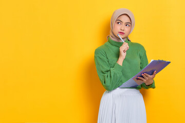 Pensive beautiful Asian woman in green sweater holding notebook looks seriously thinking about something isolated over yellow background