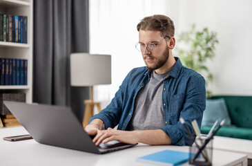 Smart young man sitting at desk and working from home on modern computer. IT specialist programmer writing complicated code on digital laptop.
