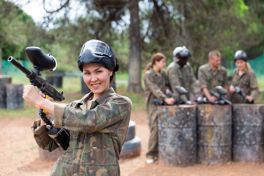 Smiling Asian Woman Paintball Player In Camouflage Posing With Gun Outdoors