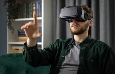 Handsome young man sitting on couch in VR headset and tapping with finger in air. Bearded guy touching virtual screen while experiencing augmented reality at home.