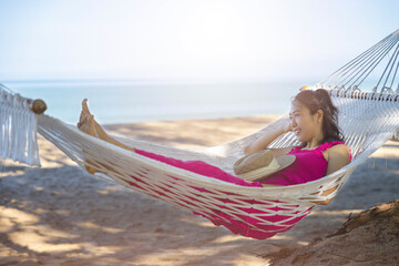Asian woman relaxing in the hammock on tropical beach, njoy her freedom and fresh air, wearing stylish hat and clothes. Happy smiling tourist in tropics in travel vacation