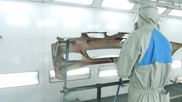 Man working in car service dressed in special overalls and respirator paints inner part of bumper red with airless spray gun in booth
