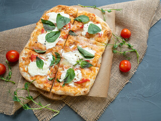 Close-up of delicious fresh crispy Roman pizza with cheese and herbs on a gray background. Fresh ripe tomatoes are nearby. Craft paper, linen napkin. Top view, flat lay