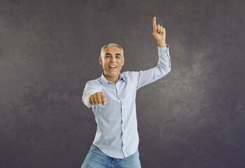 Portrait of happy energetic handsome Caucasian senior man in shirt dancing gangnam style isolated on grey background. Funny carefree modern grandad jumping, dancing, enjoying music and having fun
