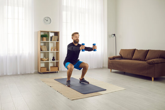 Concentrated athletic man trains squatting with dumbbells during workout at home. Beginner or amateur holds small dumbbells in outstretched hands and squats in apartment. Home sport concept.