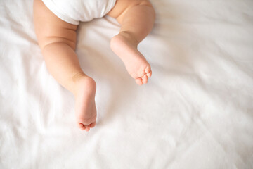 The legs of the baby in a white bodysuit on the white background of the bed. Light colors. Lifestyle.