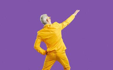Back view of funny energetic senior man in stylish outfit having fun at party. Backview of cheerful elderly guy wearing bright yellow party suit dancing isolated on solid purple colour background - Powered by Adobe