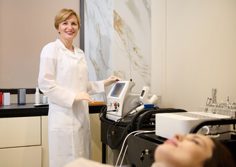 Pleasant mature woman, female beautician standing near modern medical equipment for body beauty treatment in wellness spa clinic