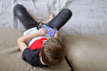Little boy playing mobile game on smartphone or watching cartoons lying on a sofa, top view. Child leisure at home, video gaming addiction