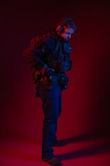 soldier in full gear with weapons. a man in headphones, body armor, with a backpack and a belt. red background. colored, blue-red light