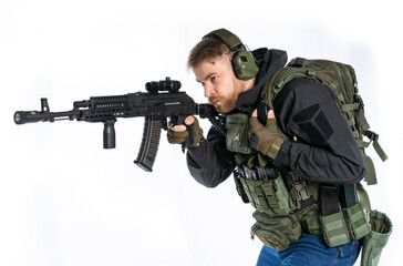 airsoft player in full gear with GG RK74 fire series guns. a man in headphones, a bulletproof vest, with a backpack and a belt, aims his machine gun to the side. profile. White background.