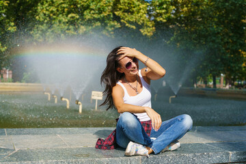 Cute caucasian woman sits by fountain in park on sunny day, enjoys the coolness from the water,...