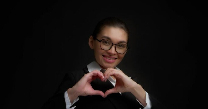 Smiling business woman shows a heart sign to the camera, expressing her love. An Asian woman in glasses and office clothes stands on an isolated black background. The concept pleasant sensations.