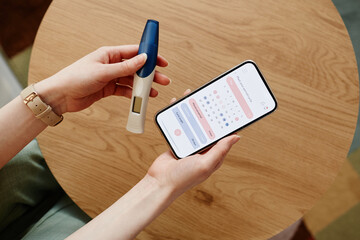 Close up of young woman calculating menstrual cycle using mobile app and holding pregnancy test - obrazy, fototapety, plakaty