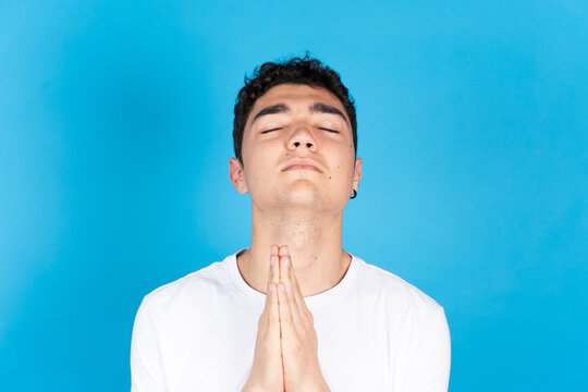 Portrait Of Hispanic Dark Teenager Boy Praying And Looking Up With Closed Eyes Isolated On Blue Background.