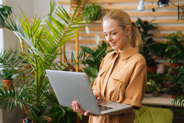 Portrait of pretty young businesswoman floral store owner standing with laptop holding in hand among green plants, looking on screen. Smiling female florist working using computer of greenhouse.