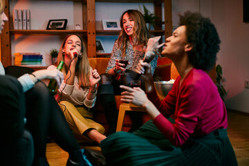 A multicultural group of female friends sings karaoke while drinking wine in the living room.