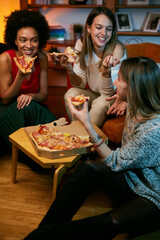 A multicultural group of cheerful young women is eating pizza while sitting in the living room. - 502330989