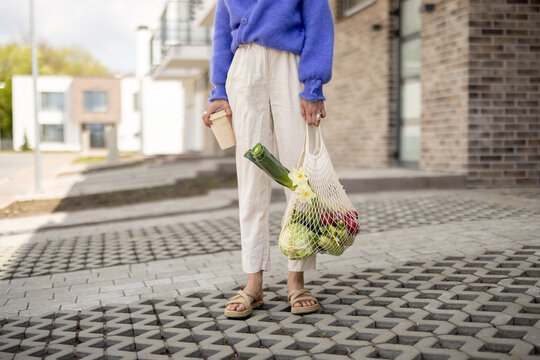 Woman holds mesh bag full of fresh vegetables and reusable coffee cup while standing at residential district, close-up. Concept of sustainability, healthy vegetarian food and eco-friendly lifestyle