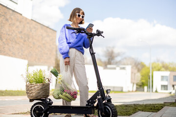 Happy stylish woman using phone while stannding with fresh vegetables and greens near electrical scooter at residential district. Concept of modern eco-friendly lifestyle and sustainability © rh2010