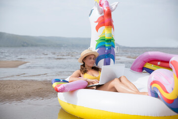 Obraz premium Working from Anywhere. Happy woman using a laptop and swim Inflatable Unicorn on a sunny day on the beach holiday 