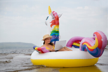 Working from Anywhere. Happy woman using a laptop and swim Inflatable Unicorn on a sunny day on the beach holiday 