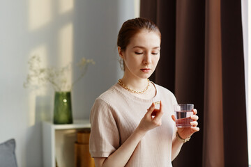 Minimal portrait of young woman holding vitamin capsule and taking supplements with glass of water at home - obrazy, fototapety, plakaty