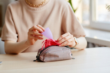 Closeup of young woman putting menstrual cup in cosmetic bag with other feminine essentials, copy...