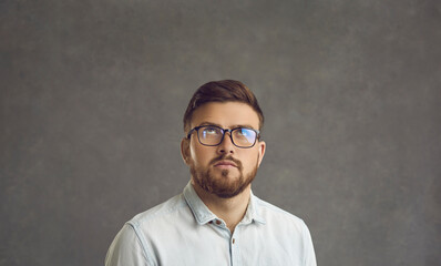 Portrait of serious handsome young man in glasses looking away imagining something or looking up at something suspicious and thinking what to do about it standing against grey studio background