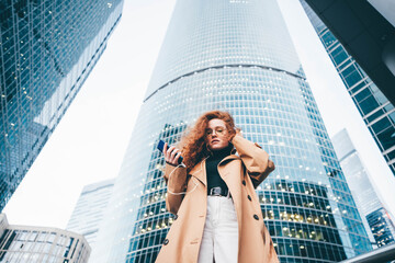 Successful woman using smartphone outdoors while standing near skyscraper.