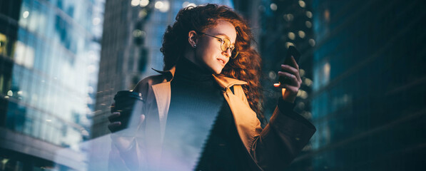 Successful woman using smartphone outdoors while standing near skyscraper at night. 