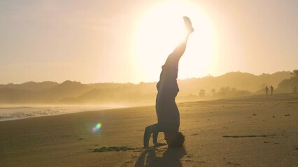 STILL SHOT: Vital young man practising yoga headstand on exotic beach at sunset. Healthy lifestyle during summer holidays in Panama. Yoga workout in golden light on a beautiful shore. - Powered by Adobe
