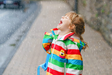 Fototapeta premium Portrait of little toddler girl on rainy day. Happy positive child running through rain, puddles. Preschool kid with rain clothes catching rain drops. Children activity on bad weather day.