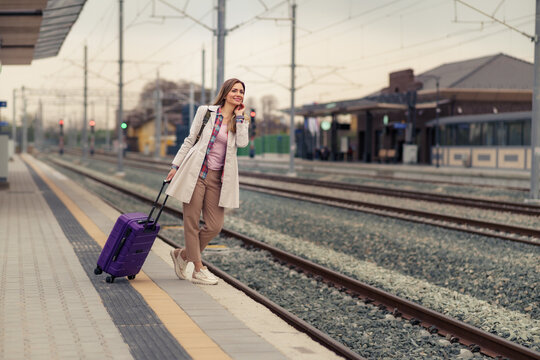 Smiling woman in 30s with suitcase waiting for her train on platform of railway station.