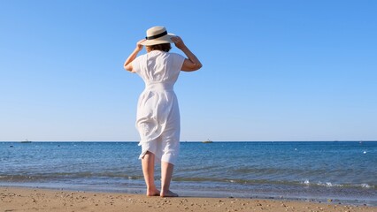 Fototapeta na wymiar back view on a mature woman in a straw hat and white dress walks along the blue sea coast on a sunny summer day, enjoying freedom and relaxation. The concept of a typical life of pensioners.