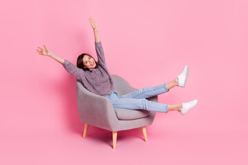 Portrait of attractive cute cheerful girl sitting in armchair rising hands up isolated over pink...