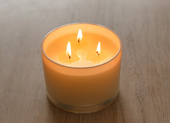 A large scented candle with three burning wicks and melting wax in a glass standing on a wooden...