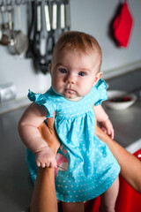 Candid portrait of little baby girl in blue dress sitting on kitchen table in mother’s hands.