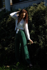 A young brunette girl in white shirt and striped trouser is stands against a green plant