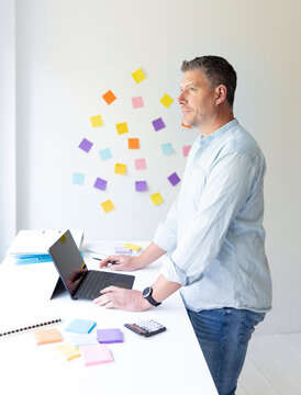 vMan with blue shirt stands at white height adjustable table and works on notebook behind him on the wall stick colorful notes