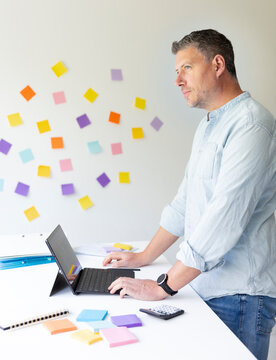 vMan with blue shirt stands at white height adjustable table and works on notebook behind him on the wall stick colorful notes