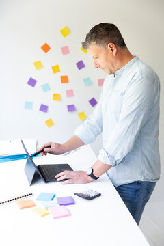 vMan with blue shirt stands at white height adjustable table and works on notebook behind him on the wall stick colorful notes