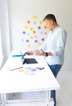 vMan with blue shirt stands at white height adjustable table and works on notebook behind him on the wall stick colorful notes