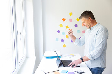 vMan with blue shirt stands at white height adjustable table and works on notebook behind him on the wall stick colorful notes