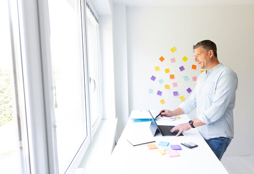 vMan with blue shirt stands at white height adjustable table and works on notebook behind him on the wall stick colorful notes