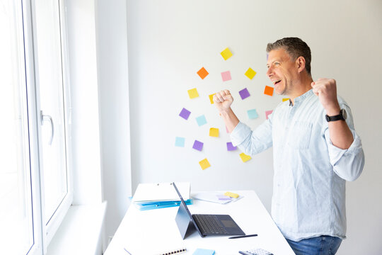 vMan with blue shirt stands at white height adjustable table and works on notebook behind him on the wall stick colorful notes