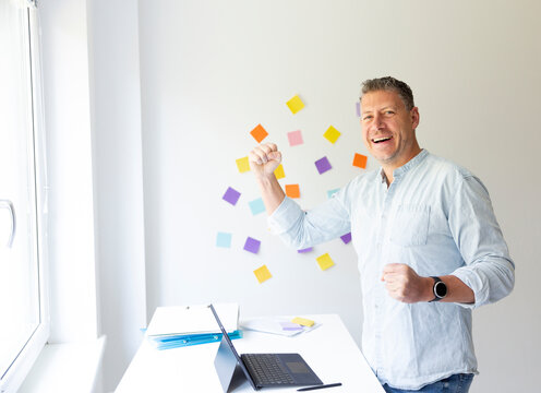 vMan with blue shirt stands at white height adjustable table and works on notebook behind him on the wall stick colorful notes