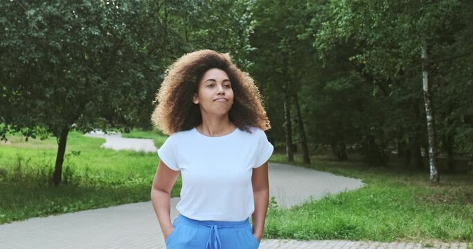Relaxed mixed race curly young modern female walking at summer forest park spending time alone outdoor. Cheerful African casual lady white t shirt resting weekend leisure activity green trees woodland